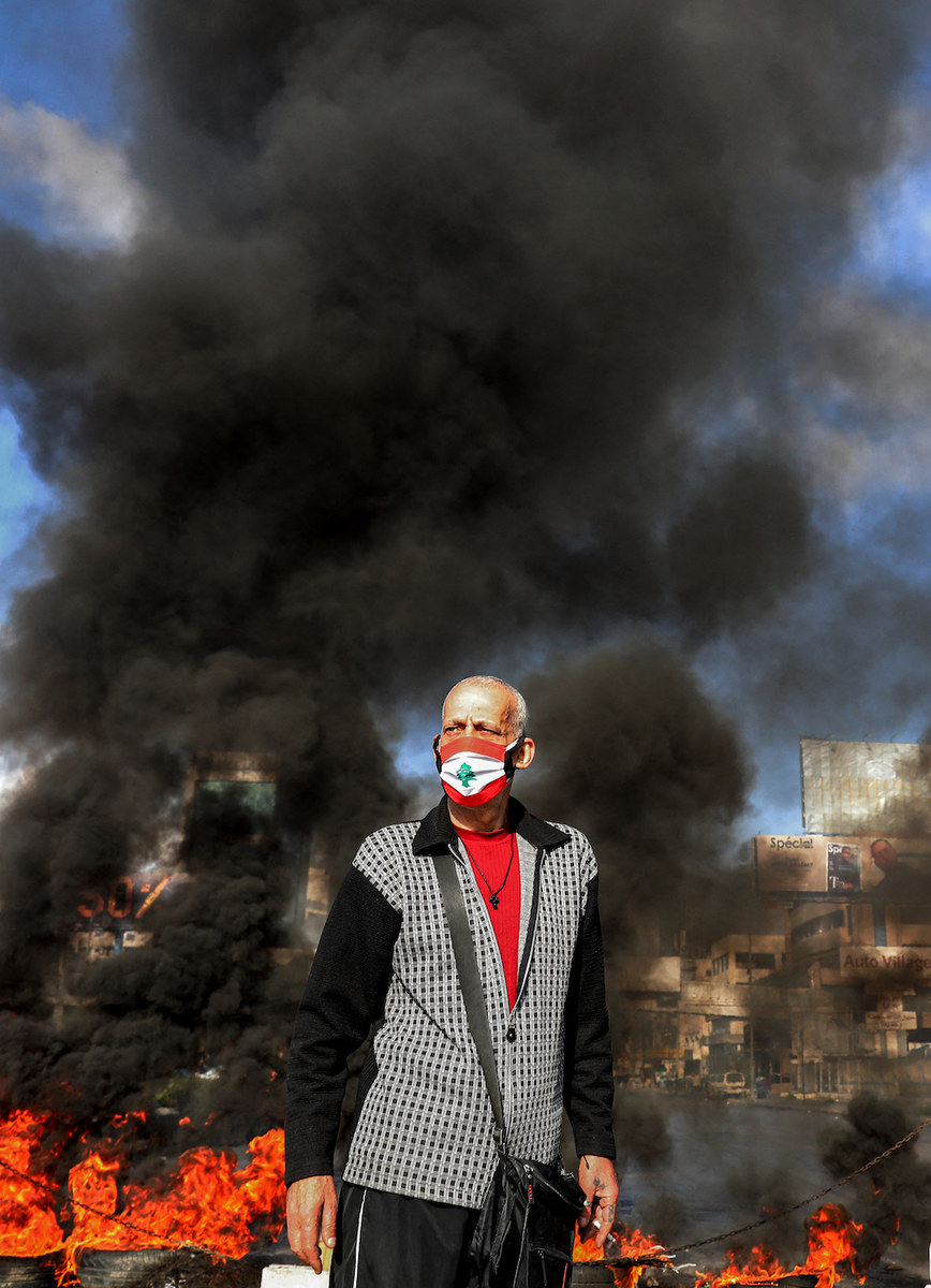Un homme portant un collier en forme de croix et vêtu d'un masque représentant le drapeau libanais se tenant à côté de pneus en flammes à un barrage routier de fortune érigé par des manifestants antigouvernementaux dans la région de Dora. (AFP/photo d'archives)